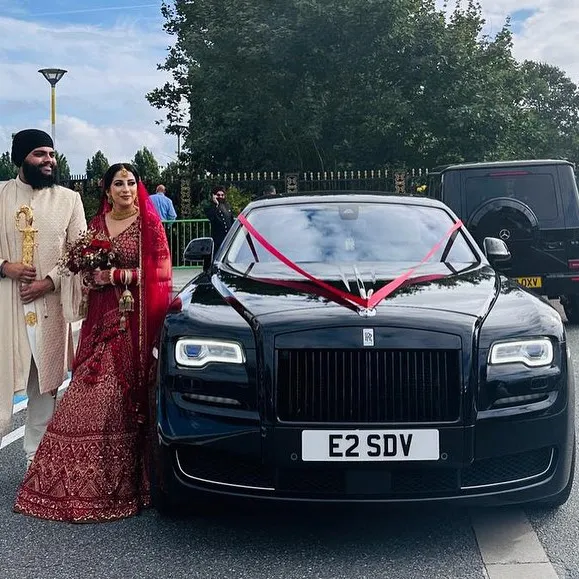 Bride and groom in traditional Sikh wedding attire standing beside a sleek black Rolls-Royce Ghost decorated with red ribbon