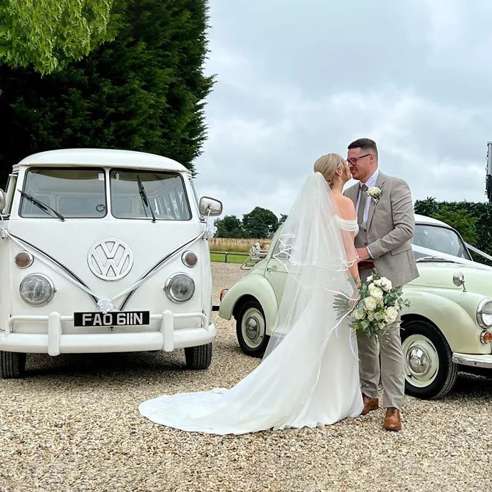 Bride and groom sharing a romantic moment between a classic white VW campervan and a soft cream Morris Minor on their wedding day.