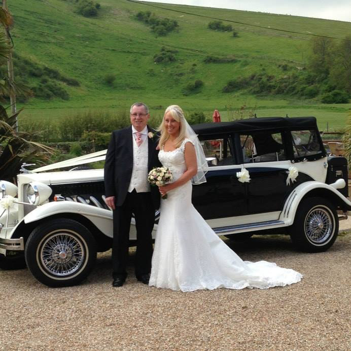 Bride in classic white gown and groom in suit posing next to a vintage-style black and white Beauford convertible at a countryside wedding.