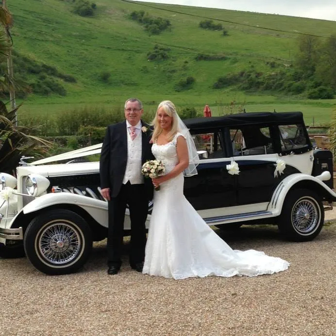 Bride in classic white gown and groom in suit posing next to a vintage-style black and white Beauford convertible at a countryside wedding.