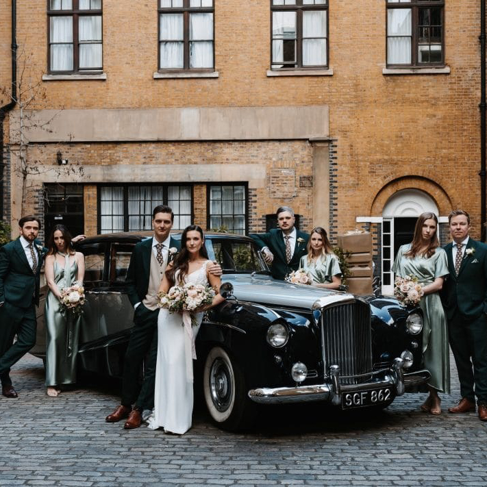 Bride and groom surrounded by bridesmaids and groomsmen in coordinated green attire, posing by a black vintage Bentley in a city courtyard.