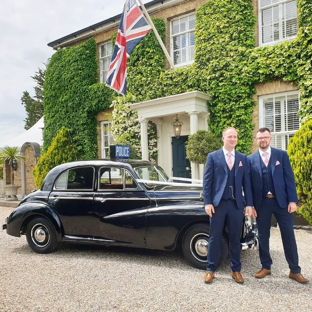Two groomsmen in navy suits posing next to a classic black Wolseley wedding car outside a traditional ivy-covered English house with Union Jack flag.