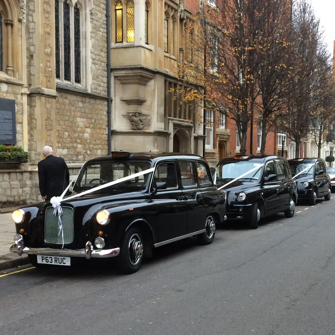 A row of classic and modern black London taxis decorated with white wedding ribbons parked outside a historic church building.