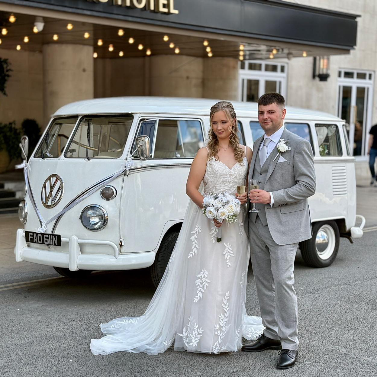 Bride in lace wedding dress and groom in grey suit posing with a retro white VW campervan parked outside a luxury hotel venue.