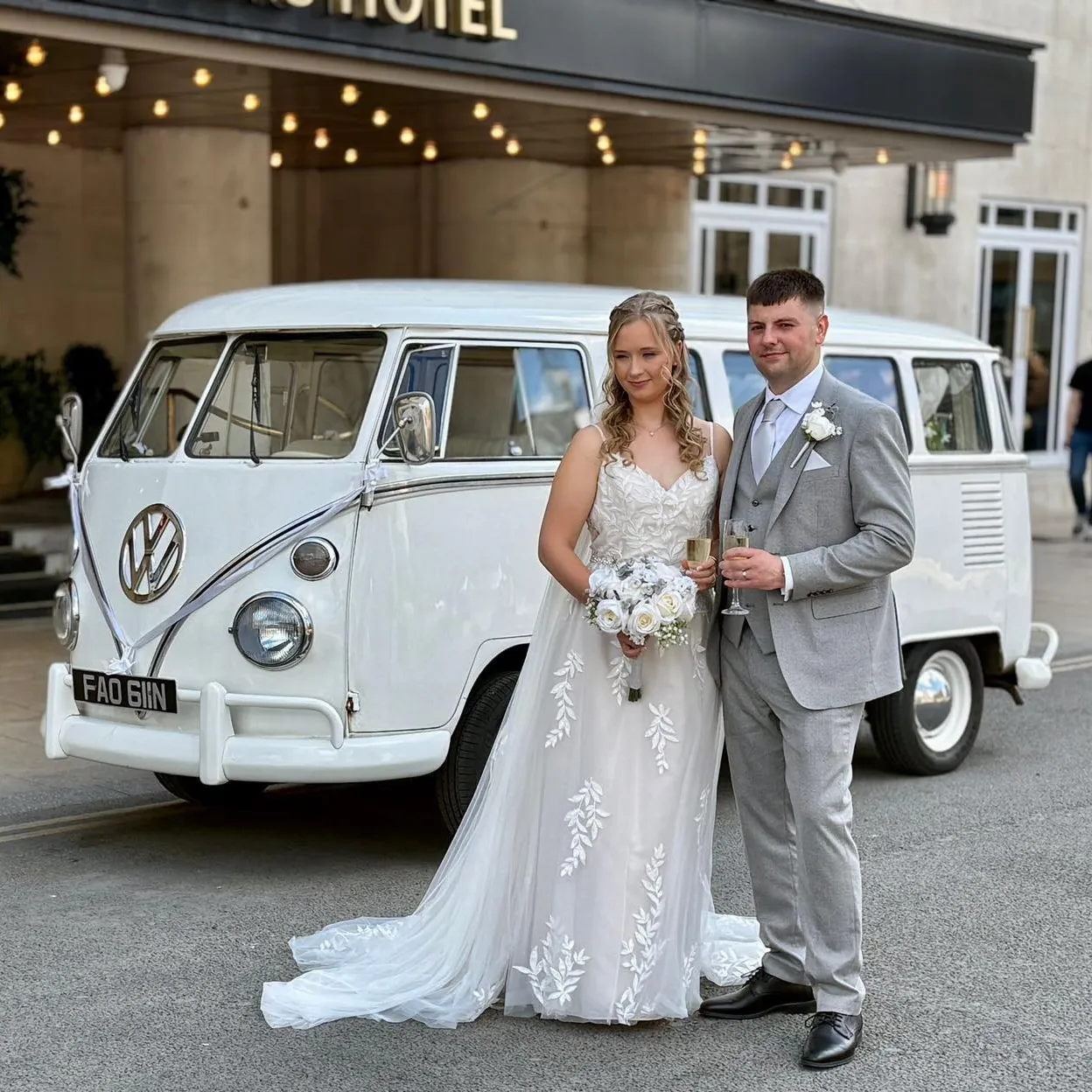 Bride in lace wedding dress and groom in grey suit posing with a retro white VW campervan parked outside a luxury hotel venue.