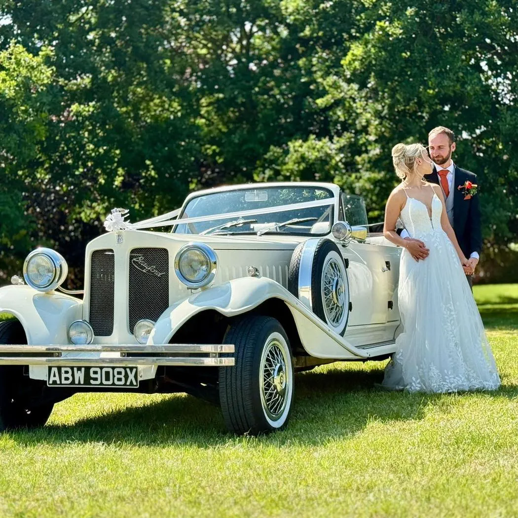 Bride and groom gazing at each other beside a white vintage Beauford convertible decorated with ribbons in a sunlit garden.