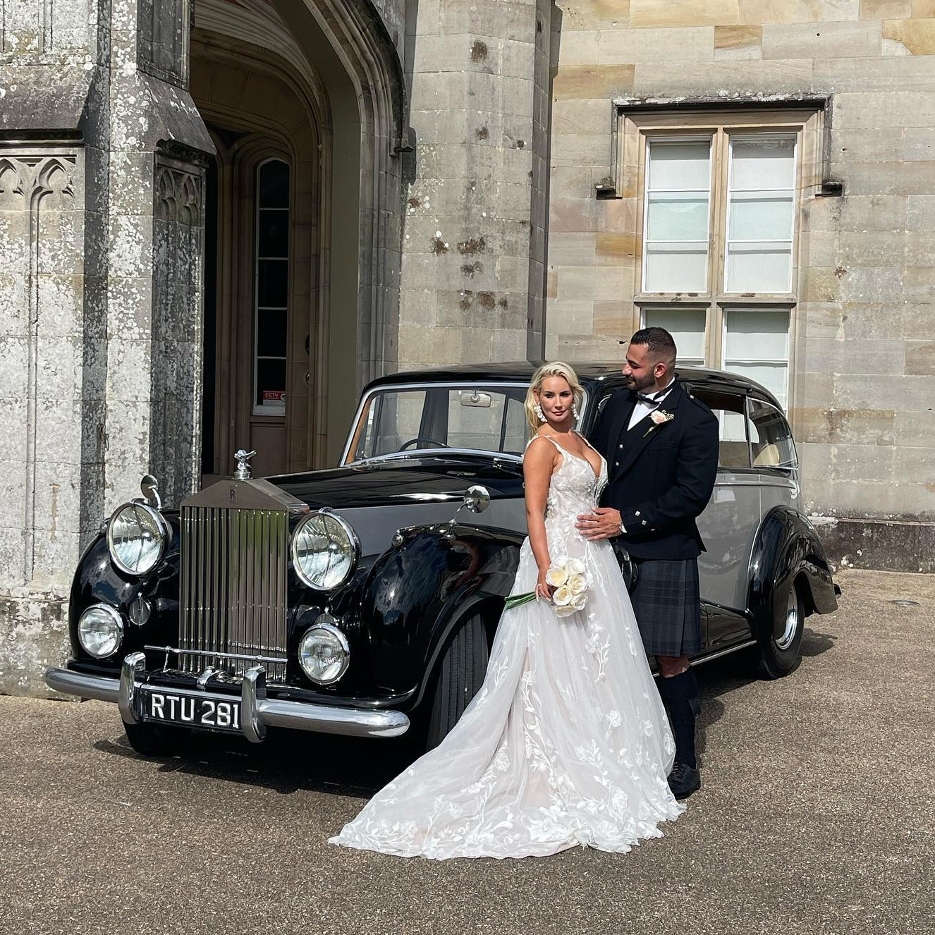 Bride in elegant lace gown and groom in traditional Scottish kilt posing in front of a vintage black and silver Rolls-Royce at a grand stone venue.