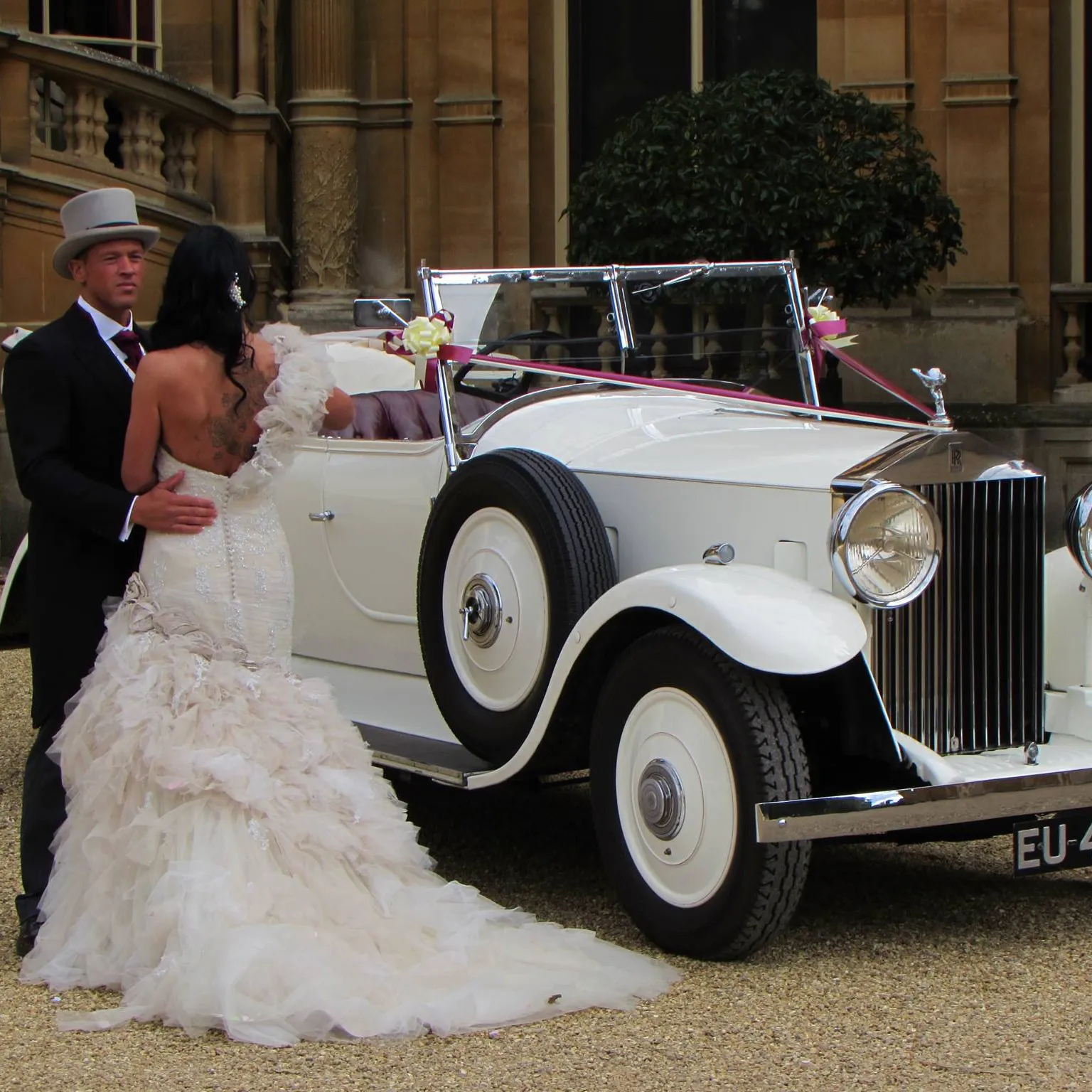 Bride in feathered gown and groom in top hat standing next to a 1930s white Rolls-Royce convertible at a grand manor house.