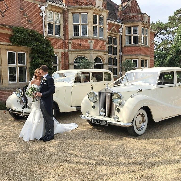 Bride and groom sharing a kiss in front of two matching vintage white Rolls-Royce wedding cars parked outside an elegant country house.