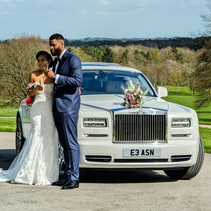 Bride and groom holding champagne flutes in front of a luxurious white Rolls-Royce Phantom adorned with fresh wedding flowers.