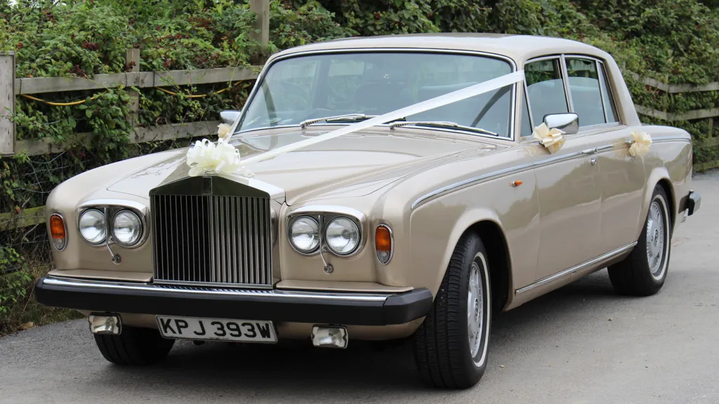 left side front view of Champagne gold coloured classic Rolls-Royce Silver Shadow dressed with wedding ribbons