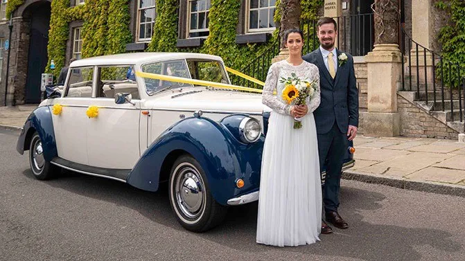 Blue and Ivory vintage-style Royale Windsor dressed with yellow ribbons and bows with Happy couple standing in front of the car. Bride is holding a bouquet of yellow sunflowers