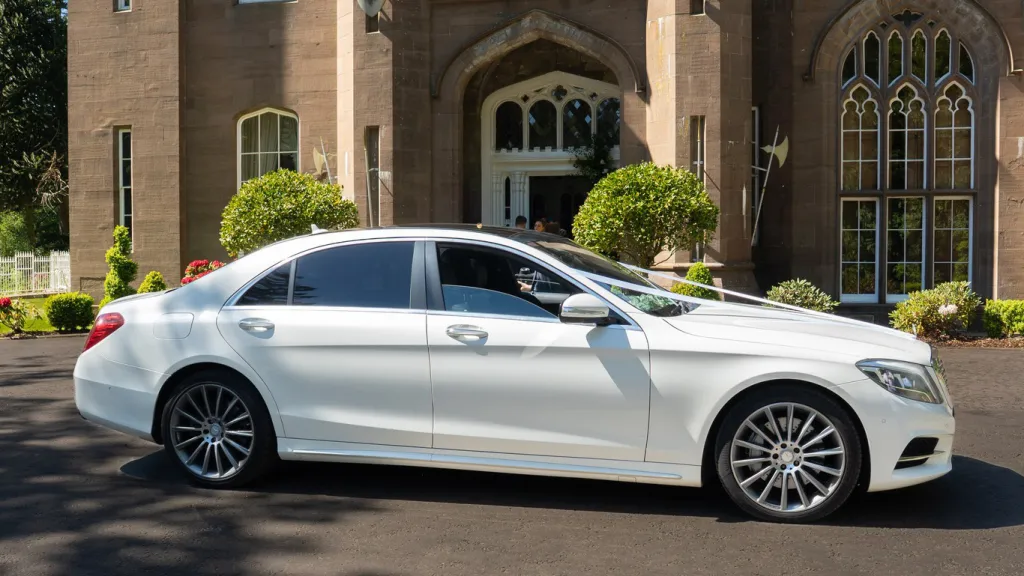 Right side view of white mercedes parked in front of a wedding venue. Car is dressed with white ribbons across the bonnet, large chrome alloy wheels