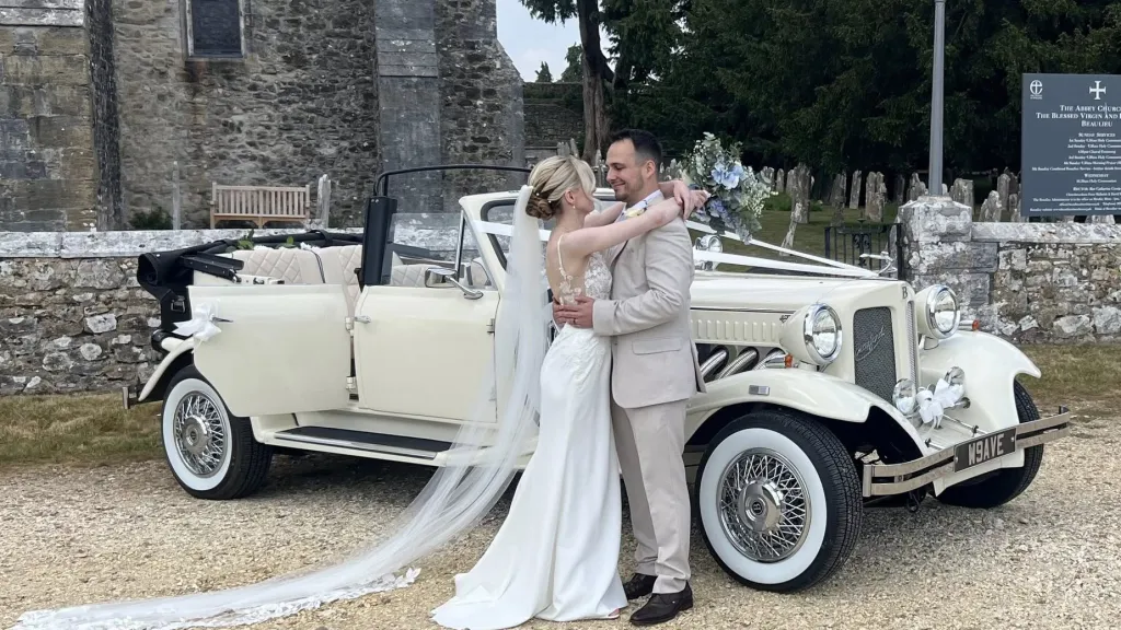 Bride and Groom holding each others and standing next to a Vintage beauford Convertible with Church in the background