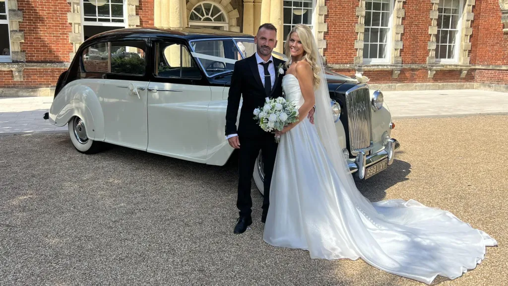 Bride in her white wedding dress holding a bouquet of flowers standing next to her groom in a black suit with a classic Austin Princess Limousine in the background