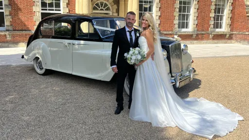 Bride in her white wedding dress holding a bouquet of flowers standing next to her groom in a black suit with a classic Austin Princess Limousine in the background
