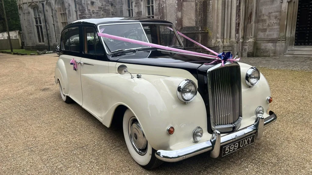 Austin Princess Limousine in Ivory with Black roof and bonnet dressed with a pink ribbon parked on a gravel parking in front of wedding venue