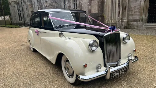 Austin Princess Limousine in Ivory with Black roof and bonnet dressed with a pink ribbon parked on a gravel parking in front of wedding venue