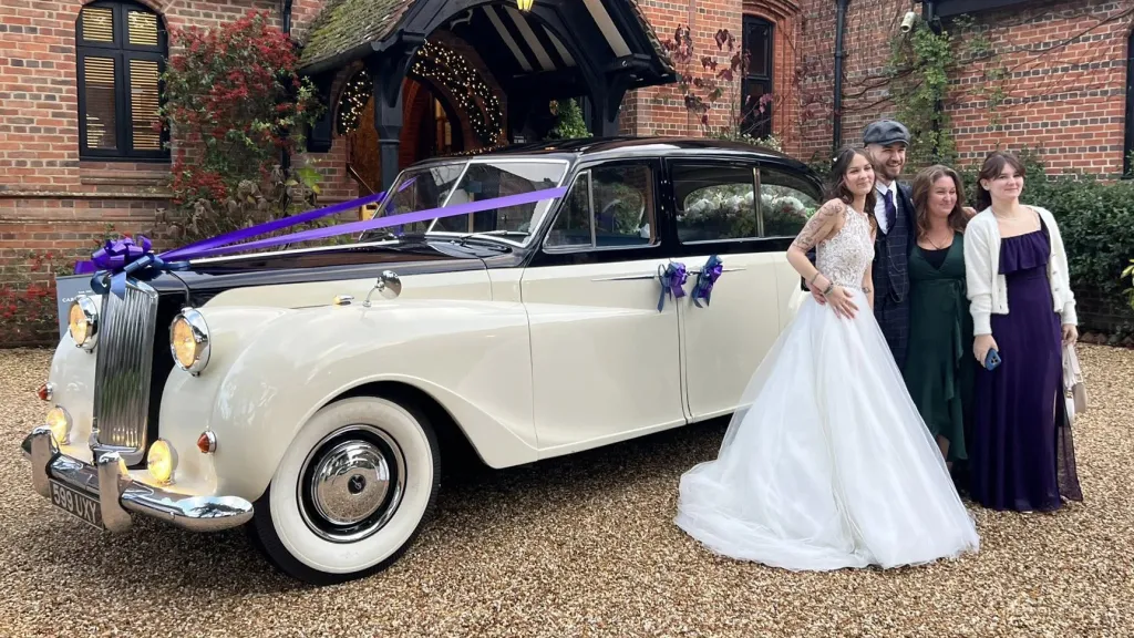 Bride with wedding guests standing in front of a classic Austin Princess Limousine in Ivory with cadbury purple ribbons