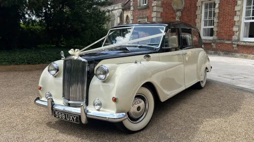 Austin Princess Limousine in Ivory with Black bonnet and roof decorated with Ivory Ribbons and bows, large white wall tires, chrome front bumper. Parked in front of wedding venue