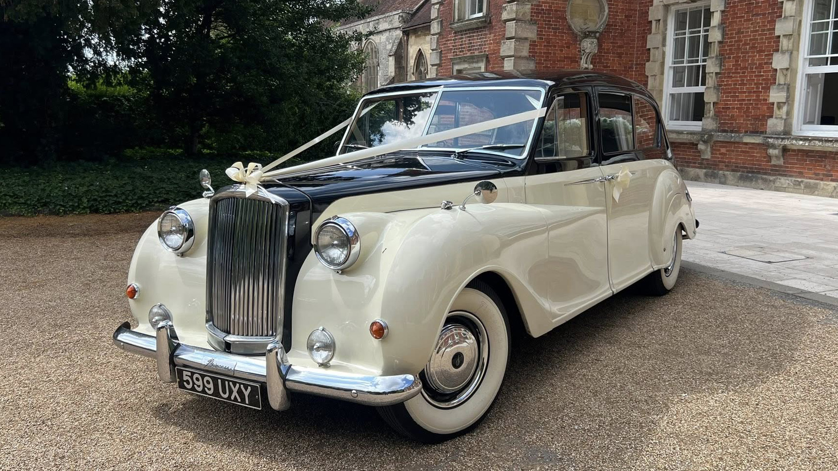 Austin Princess Limousine in Ivory with Black bonnet and roof decorated with Ivory Ribbons and bows, large white wall tires, chrome front bumper. Parked in front of wedding venue