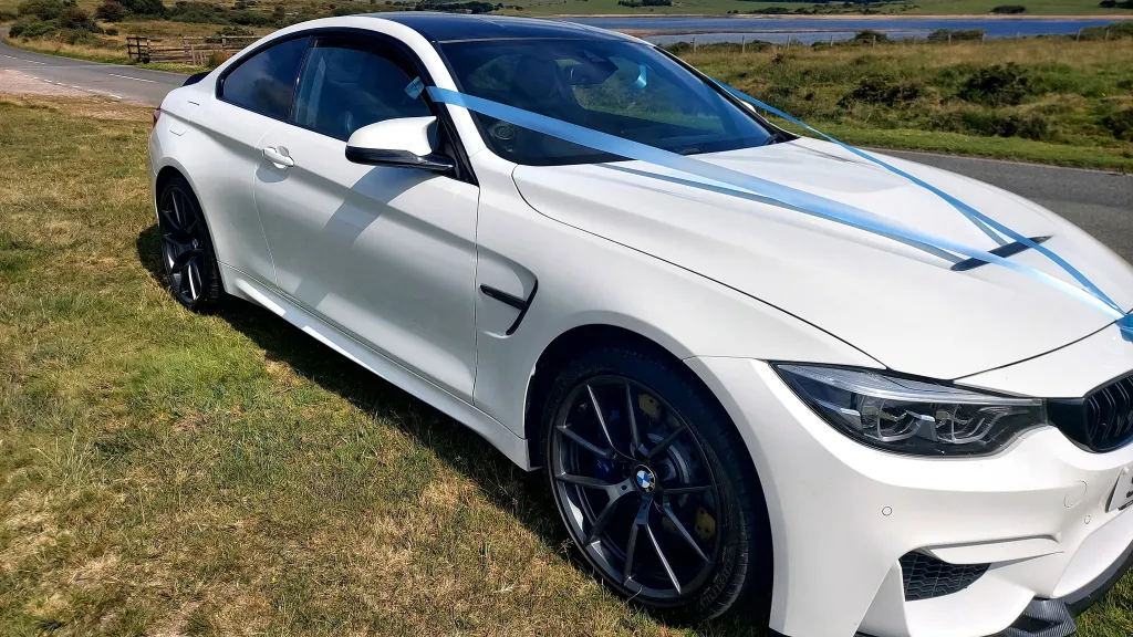 Front right side view of white BMW with large black alloy wheels and baby blue ribbons across the front bonnet