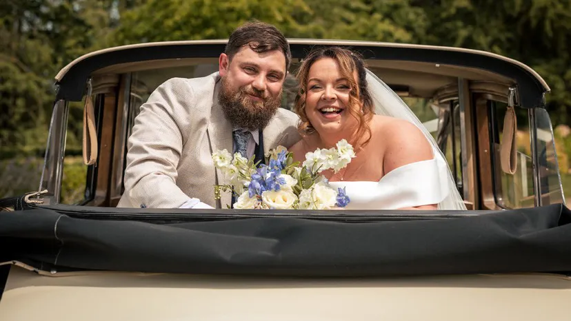 Smiling Groom with Bride holding a bouquet of flowers in the back of a Convertible classic limousine looking backward.