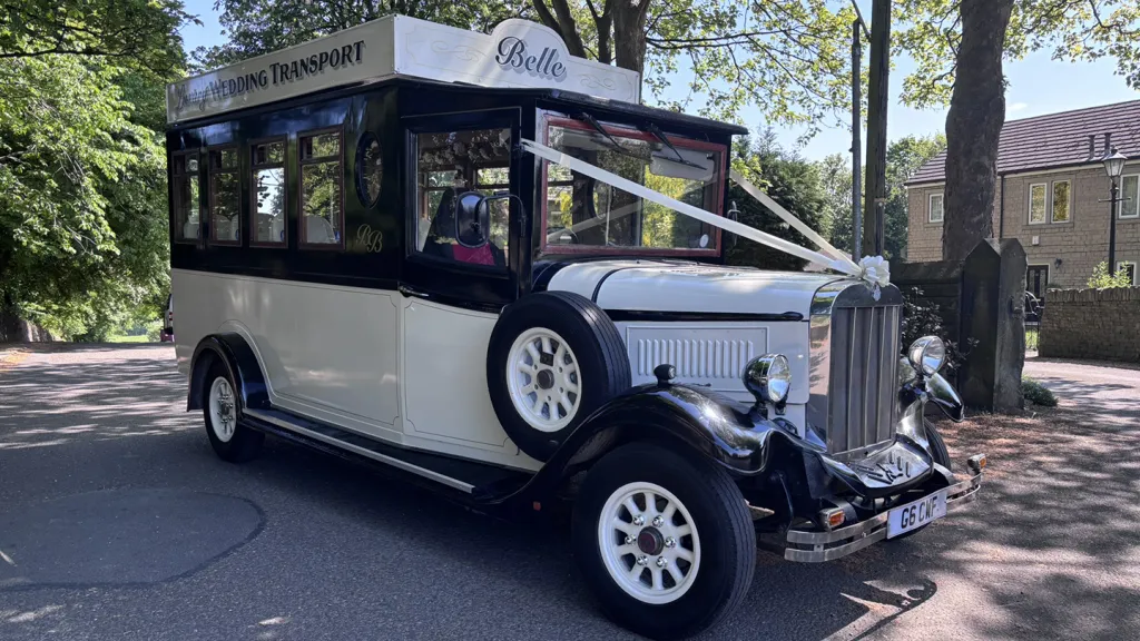Asquith Bus in Black and Ivory dressed with wjhite wedding ribbon and bow across front bonnet. Spare whelel mounted on the right side of the bus and a l;arge decorative sign at the top with wording "Belle"