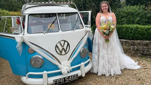 Classic VW campervan decorated with large white ribbons and bows with a Bride standing next to the camper holding a bouquet of flowers