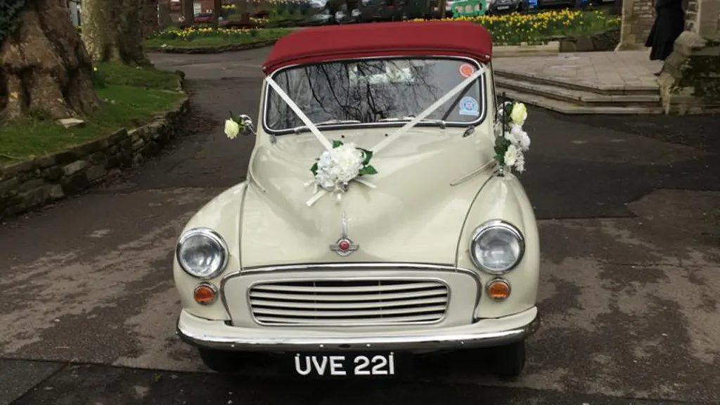 Full front view of Ivory classic Morris Minor Convertible with convertible soft top roof closed and wedding ribbons and flowers decorating the car's bonnet