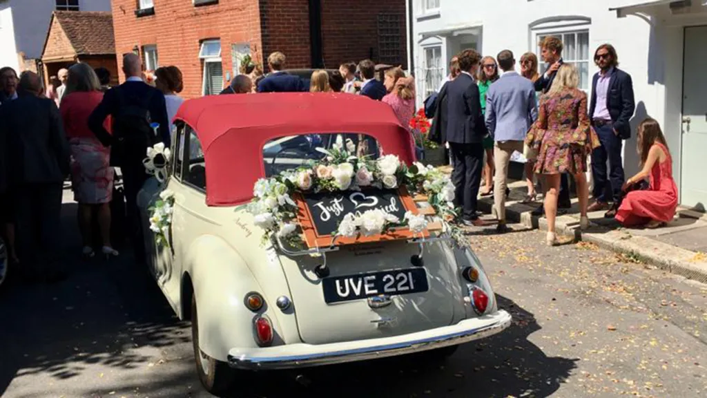 Classic Morris Minor Convertible with burgundy roof closed, flower decorated with Just Married sign at the rear of the vehicle. Wedding Guests can be seen in the background