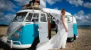 Smiling Bride in her white dress standing in front of a Blue & White Classic VW campervan decorated with white wedding ribbons