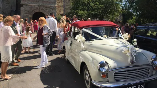 Front view of classic Morris Minor with convertible burgundy sift top roof closed parked in front of a Church with wedding guests in the background