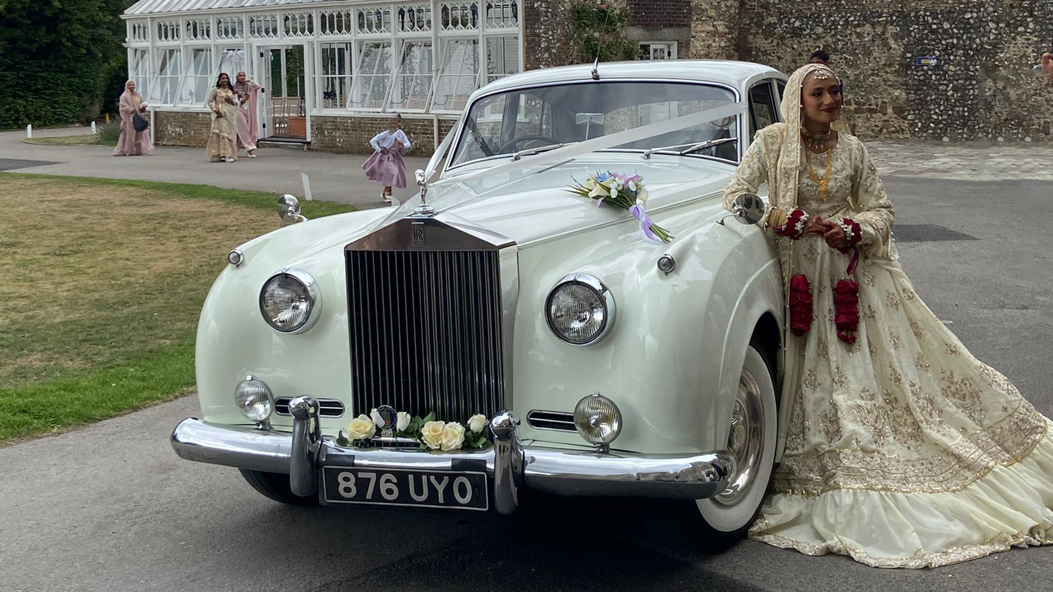Asian bride in a gold wedding dress leaning on a white classic Rolls-Royce Silver Cloud