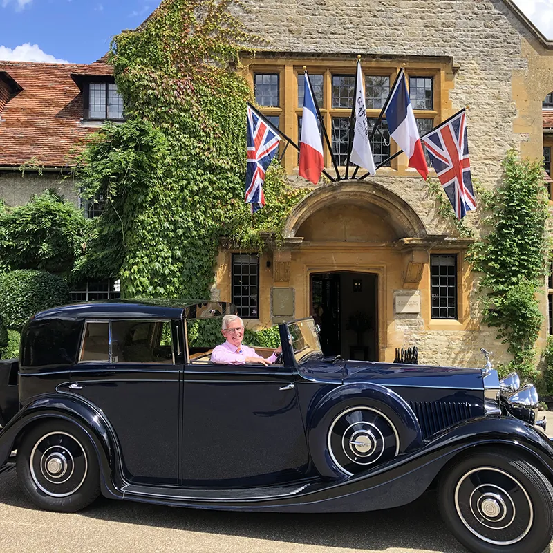A navy blue vintage Rolls-Royce parked outside a country house with flags, chauffeur at the wheel, ready for the bride and groom.