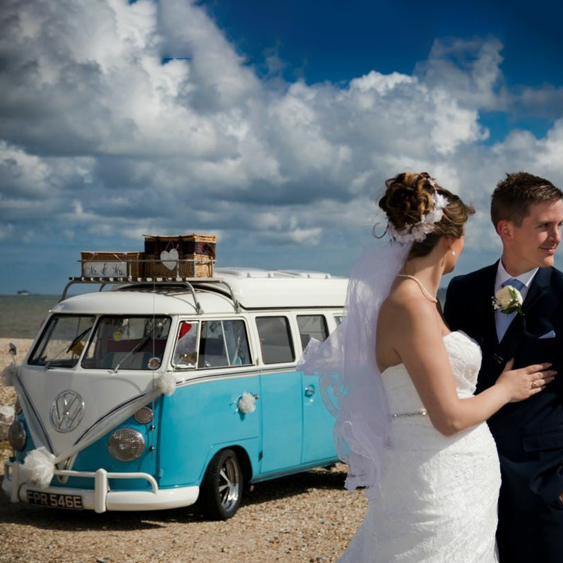 A retro blue and white VW campervan as quirky wedding transport, with the bride and groom standing together in front of it.