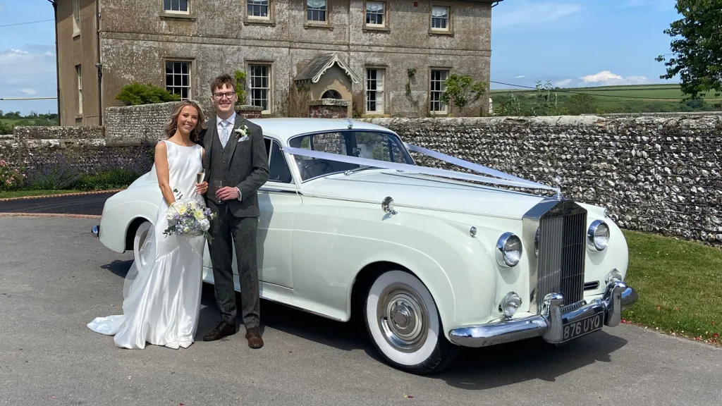 Bride and groom smiling in front of a white Rolls-Royce Silver Cloud outside a manor house