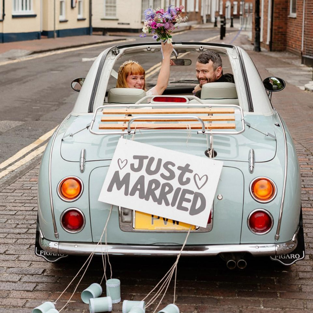 Newlyweds smiling and waving from the back of a pale blue Fiat convertible decorated with a “Just Married” sign.