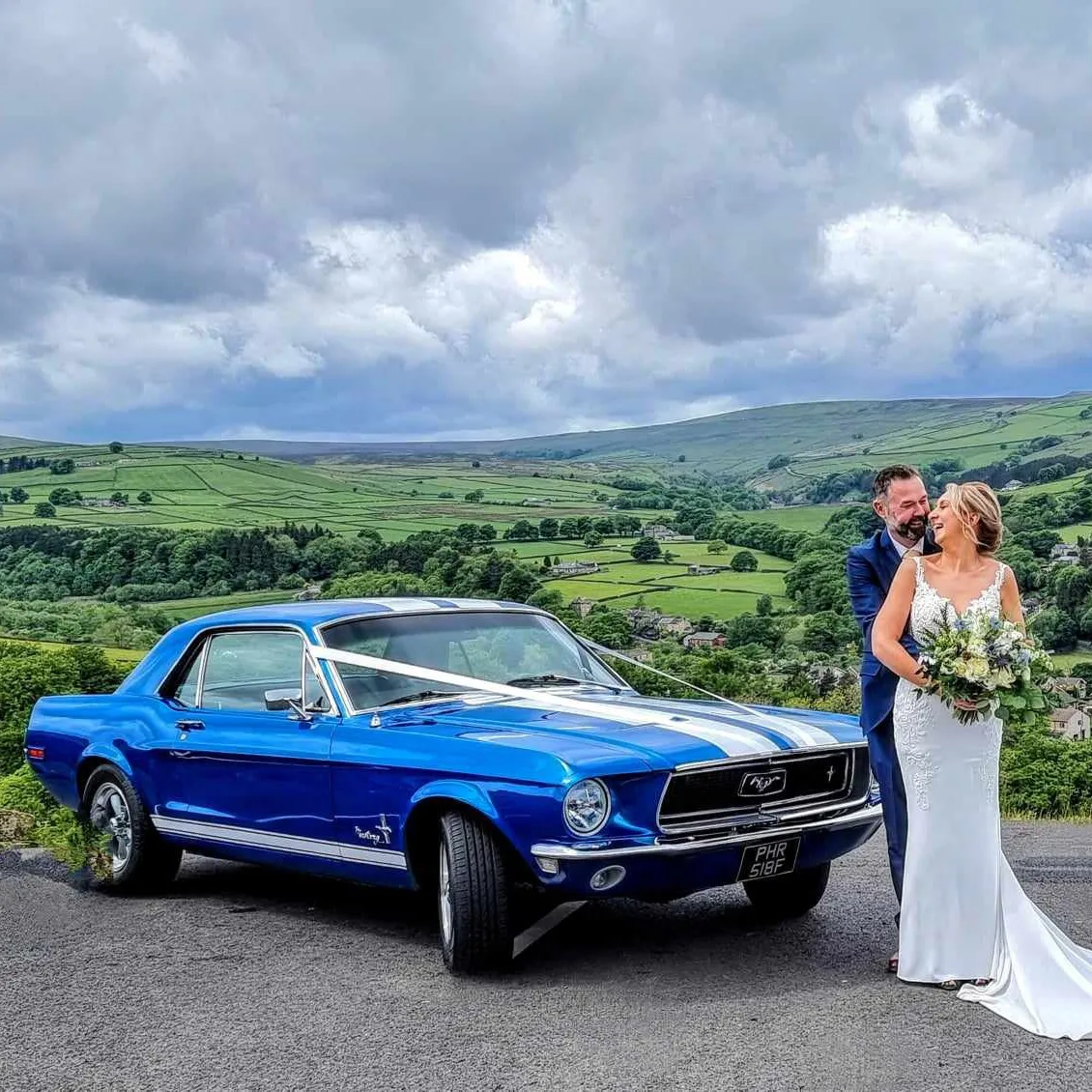 A striking electric blue Ford Mustang parked on a hilltop, with the bride and groom posing against a scenic countryside backdrop.