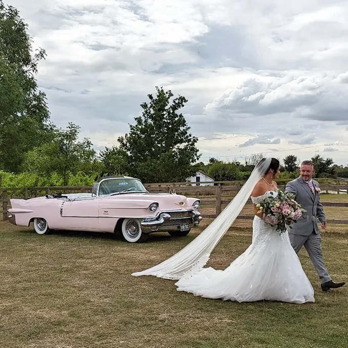 Newlyweds walking hand in hand with a classic pale pink Cadillac parked nearby on a rustic countryside setting.