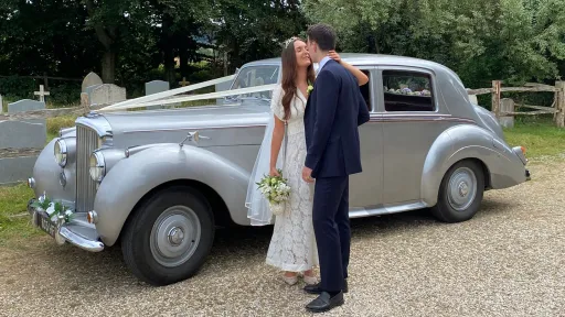 Bride and groom facing each other beside a silver Bentley R-Type wedding car