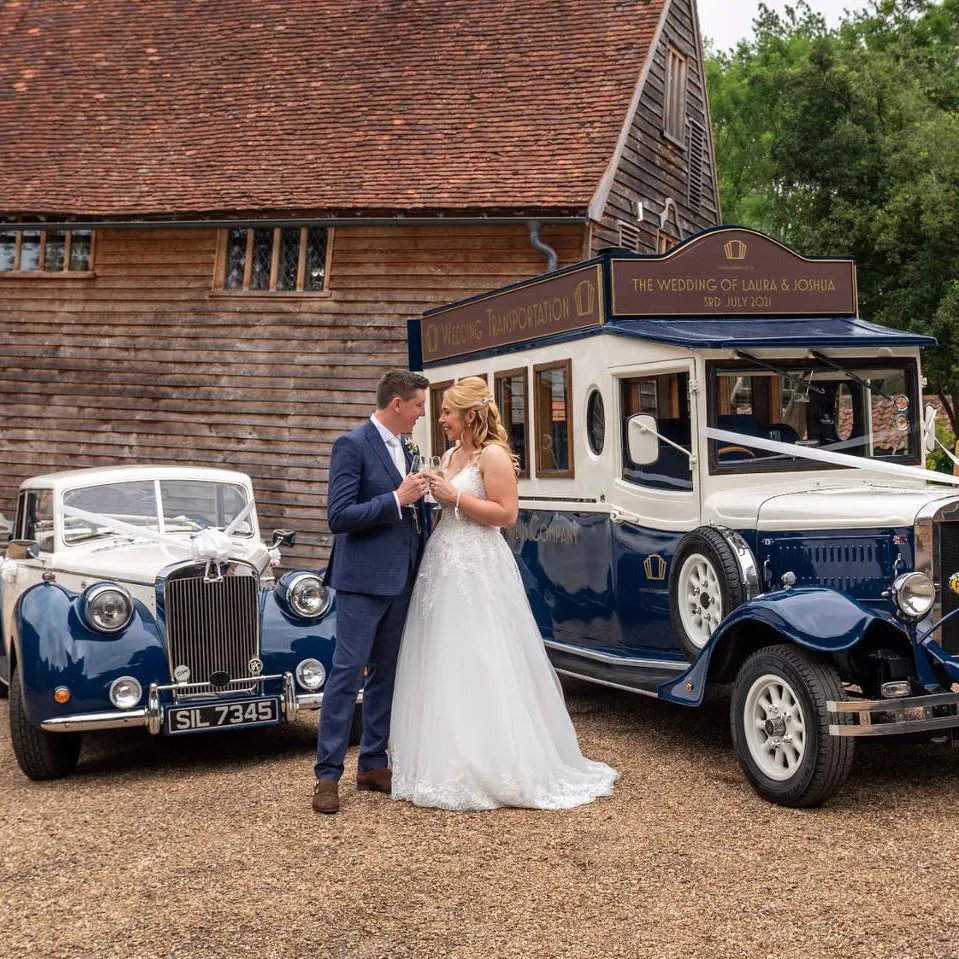 A couple embracing beside two vintage wedding cars, including a blue classic bus alongside a cream and blue convertible car, outside a rustic barn.