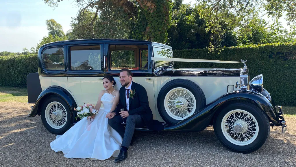 Bride and groom sitting on grass with a vintage Rolls-Royce 20/25 in the background