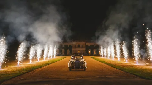 White Rolls-Royce Silver Cloud driving up an estate driveway at night with fireworks flaring on both sides