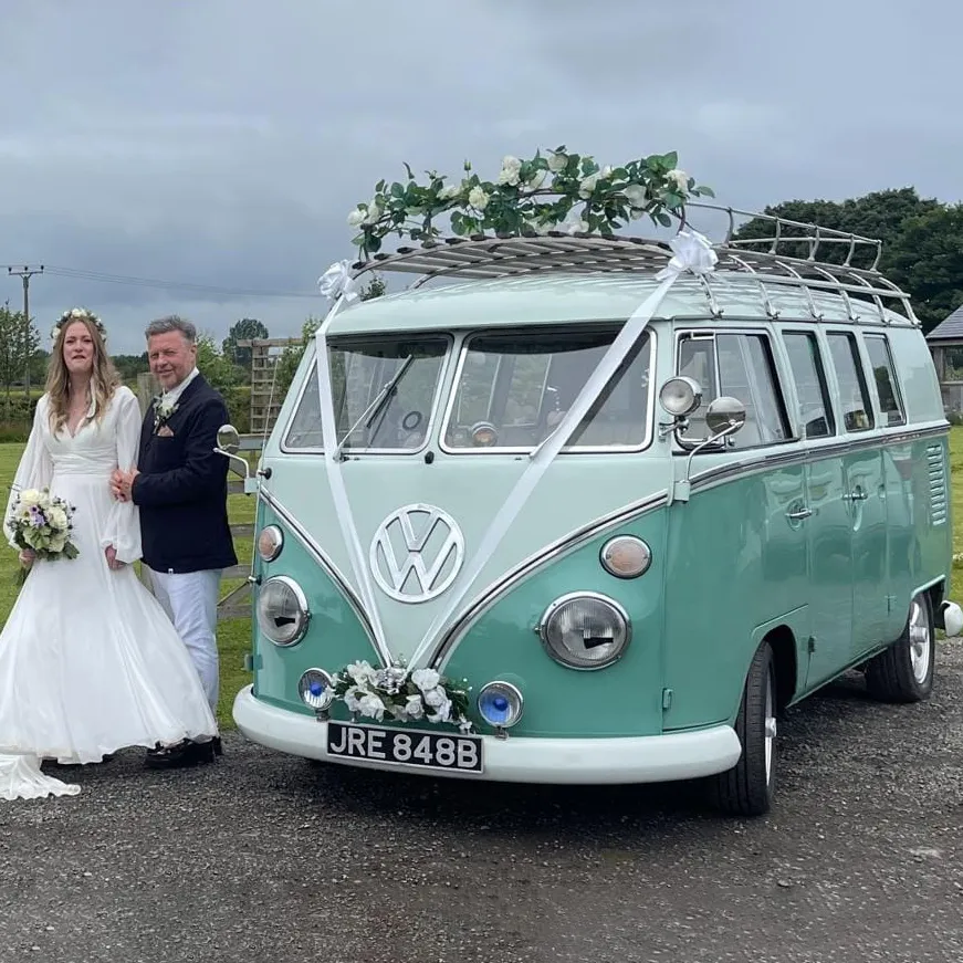 Bride and groom standing beside a mint green Volkswagen splitscreen Campervan decorated with white flowers and green foliage