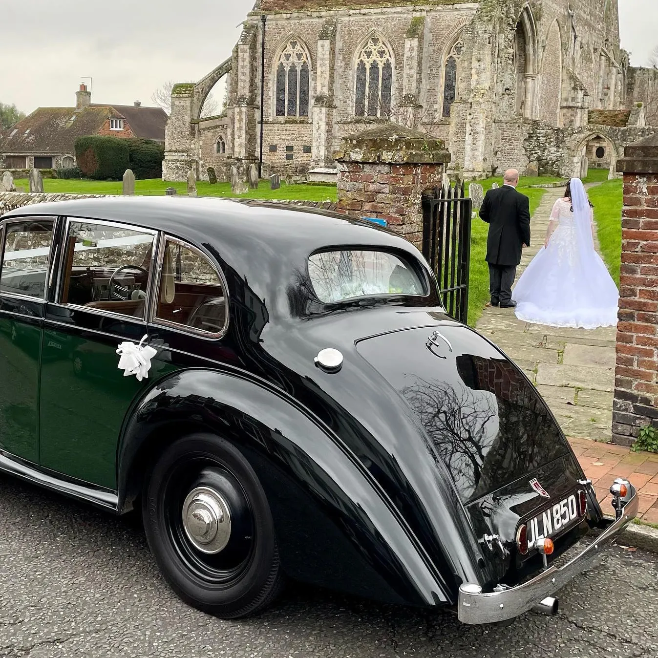 Bride and groom walking toward a church in the background with a dark green Lancaster classic car