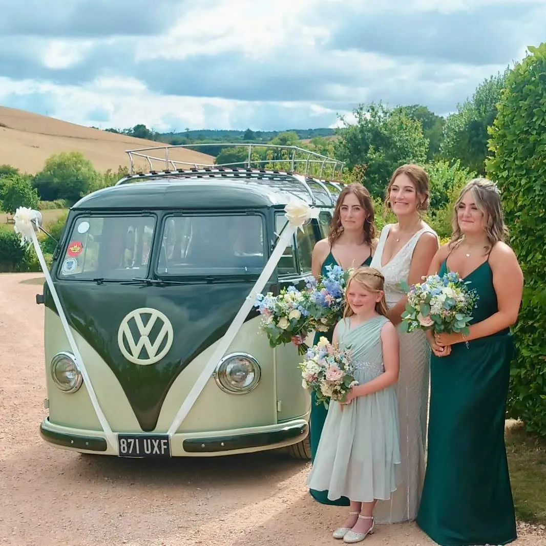 Bride and bridesmaids in green dresses standing beside a two-tone green Volkswagen splitscreen Campervan decorated with white ribbon