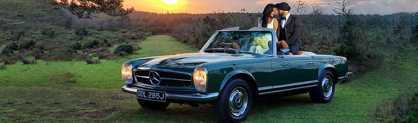 Bride and groom in a classic dark green Mercedes convertible at sunset, parked on a scenic countryside field, creating a romantic wedding moment.