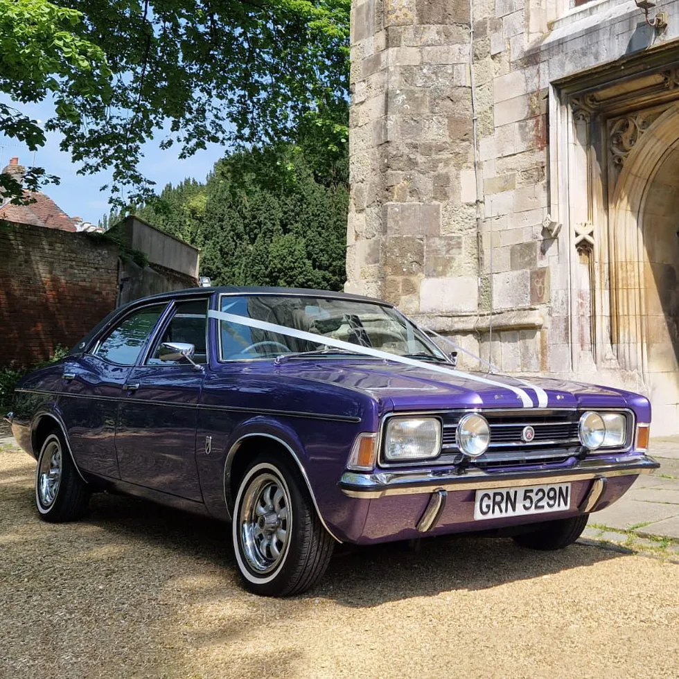 A vintage purple classic car decorated with ivory ribbons parked outside a stone church, ready for the bride and groom.