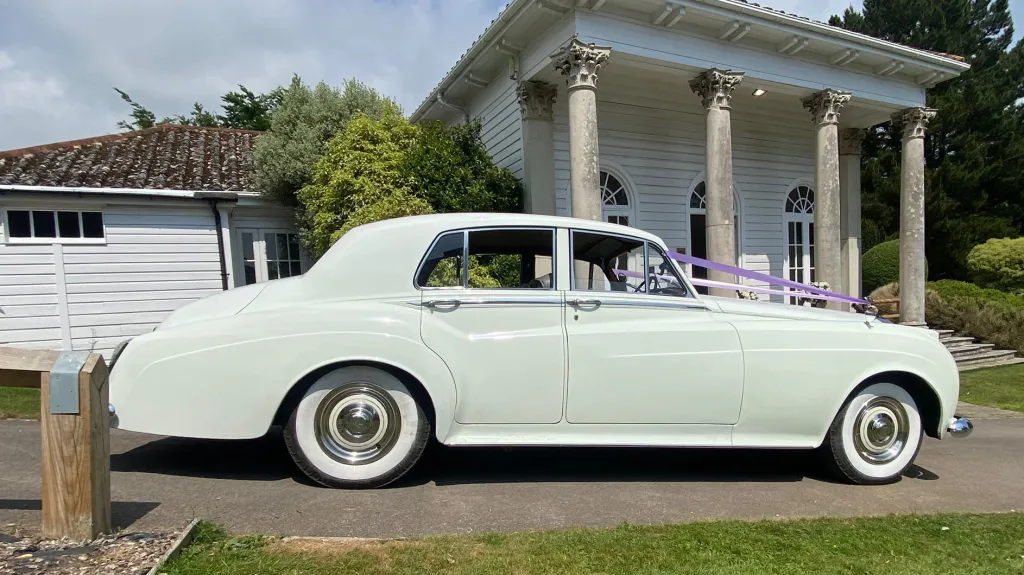 Right side profile of a white Rolls-Royce Silver Cloud parked outside a columned house